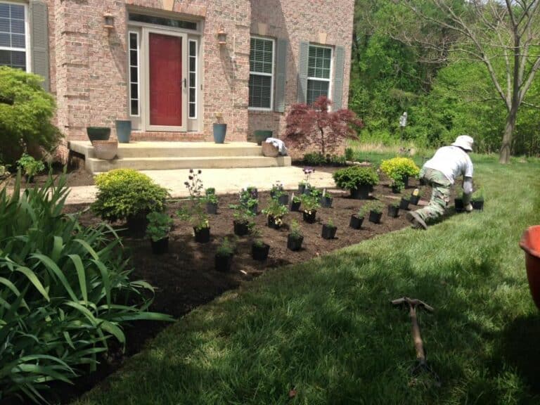 Lush backyard garden with new flower beds and landscaping, featuring a brick house exterior, potted plants, and a landscaper planting flowers in a vibrant outdoor space.
