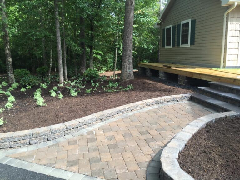 Natural stone and brick paver outdoor patio with landscaping, wooden deck, and lush greenery, ideal for outdoor living spaces, hosting, and relaxation. Perfect for enhancing your backyard with durable, stylish design.