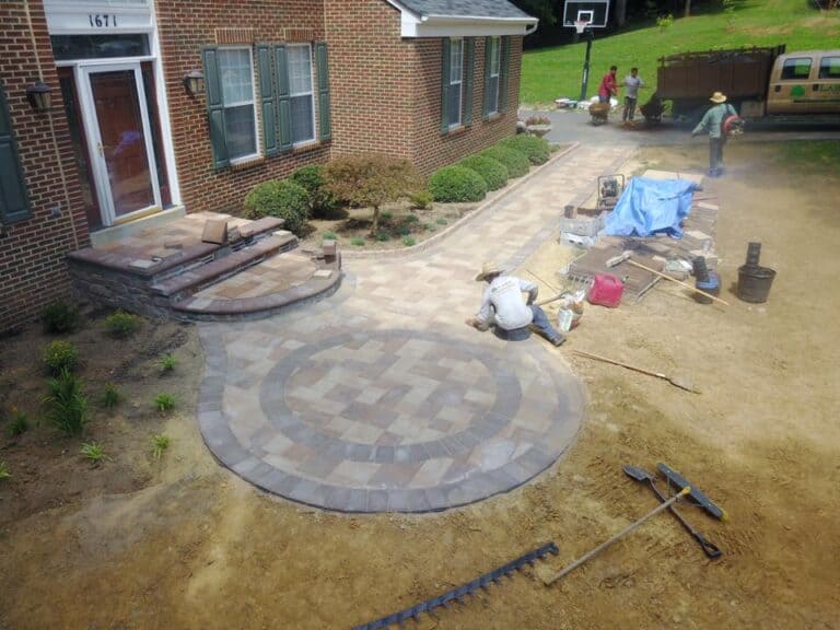 Multicolored paver patio installation in progress outside brick house with landscaping and workers constructing outdoor living space.
