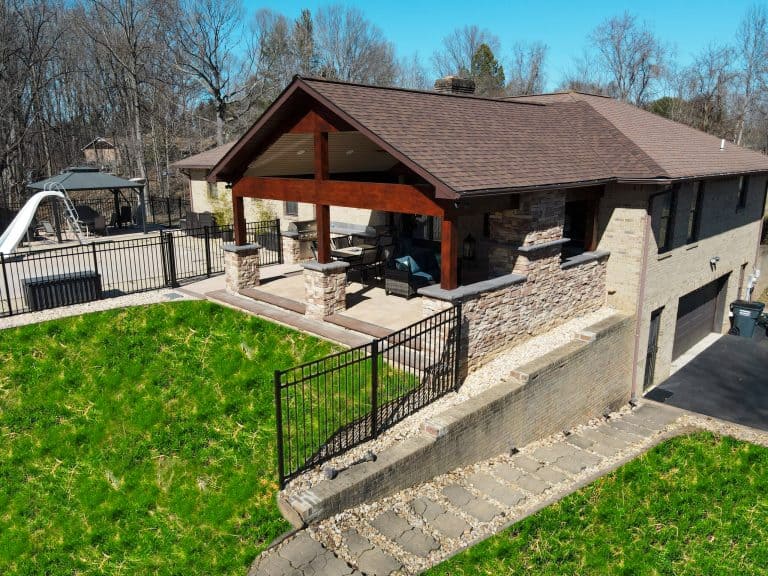 Spacious outdoor living space with a covered patio, stone fireplace, modern seating area, and a fenced backyard featuring a lush green lawn and a play area, perfect for relaxing and entertaining.