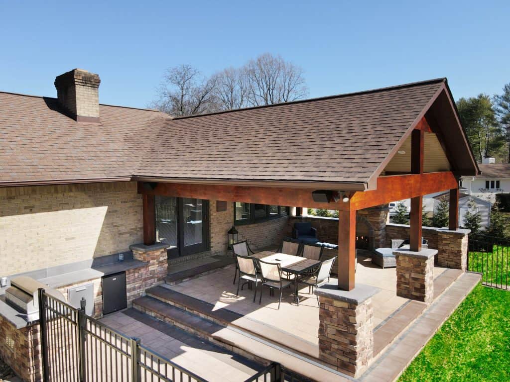 Elegant outdoor covered patio with stone columns, wooden beams, and a cozy fireplace, perfect for relaxing and entertaining in a beautiful backyard setting.