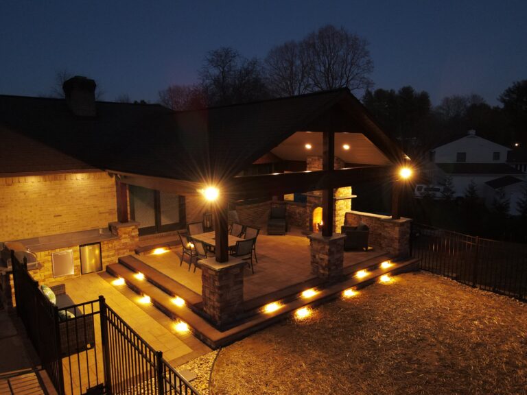 Spacious outdoor living space with a covered patio, cozy fireplace, and stylish lighting, perfect for relaxing and entertaining during evenings in a beautifully designed backyard.