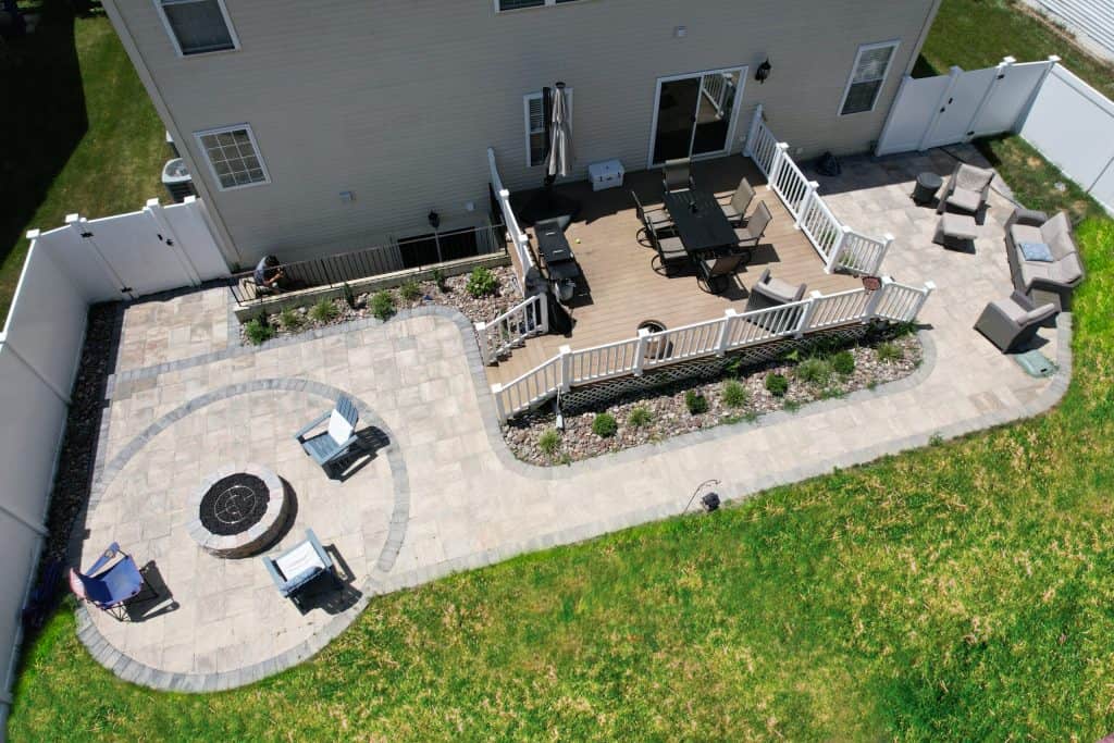Bright outdoor living space featuring a spacious patio, fire pit area, seating, and a dining deck, ideal for entertaining and relaxation. Perfect for custom outdoor living spaces and yard upgrades.