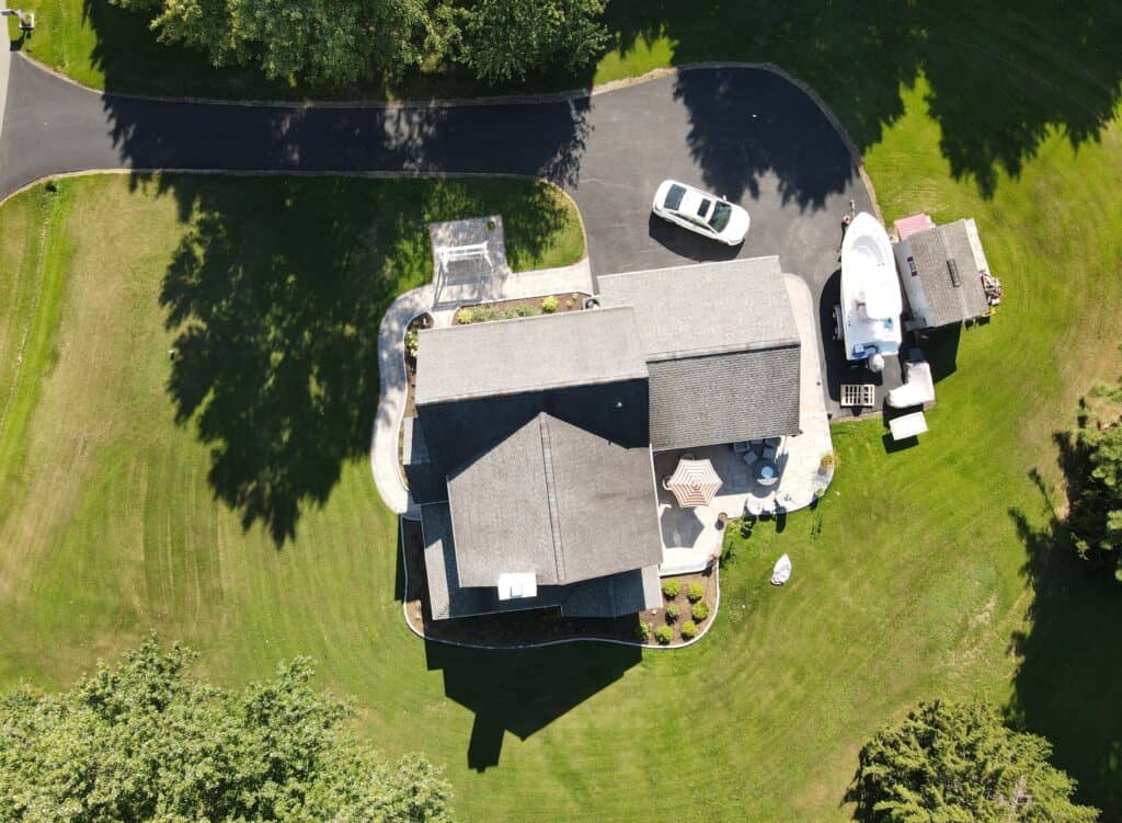 Aerial view of a landscaped backyard featuring a patio, outdoor living space with seating, lush green lawn, mature trees, and a driveway with a car and boat trailer, perfect for outdoor entertaining and relaxing.
