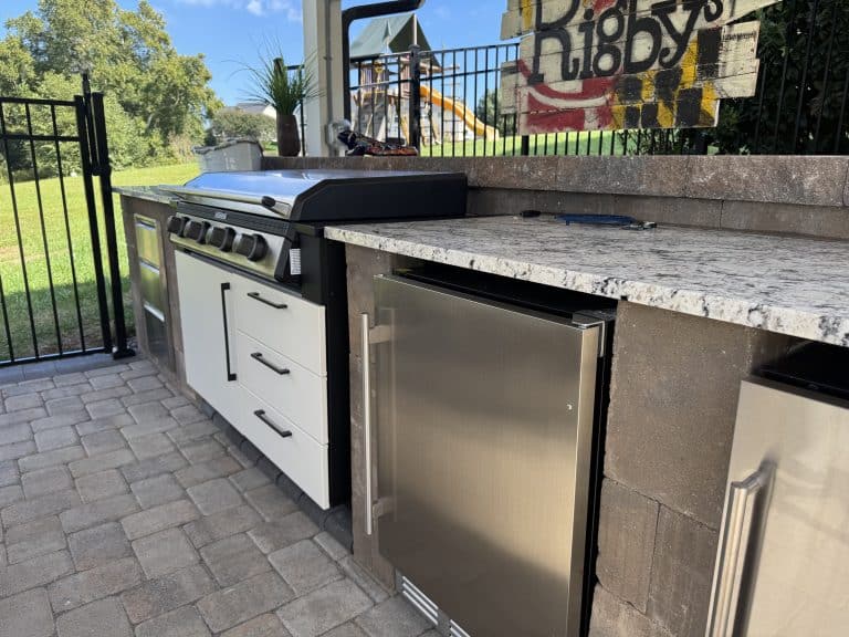 Outdoor kitchen with built-in grill, mini fridge, and granite countertops in a landscaped backyard featuring a play area and lush greenery. Perfect for outdoor entertaining and cooking.