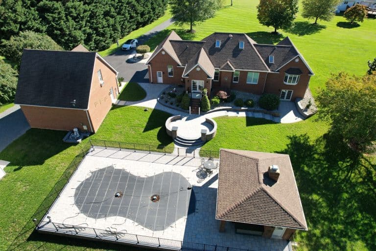 A spacious backyard featuring a custom-designed swimming pool, lush green lawn, and a large brick house with a patio area for outdoor entertaining.