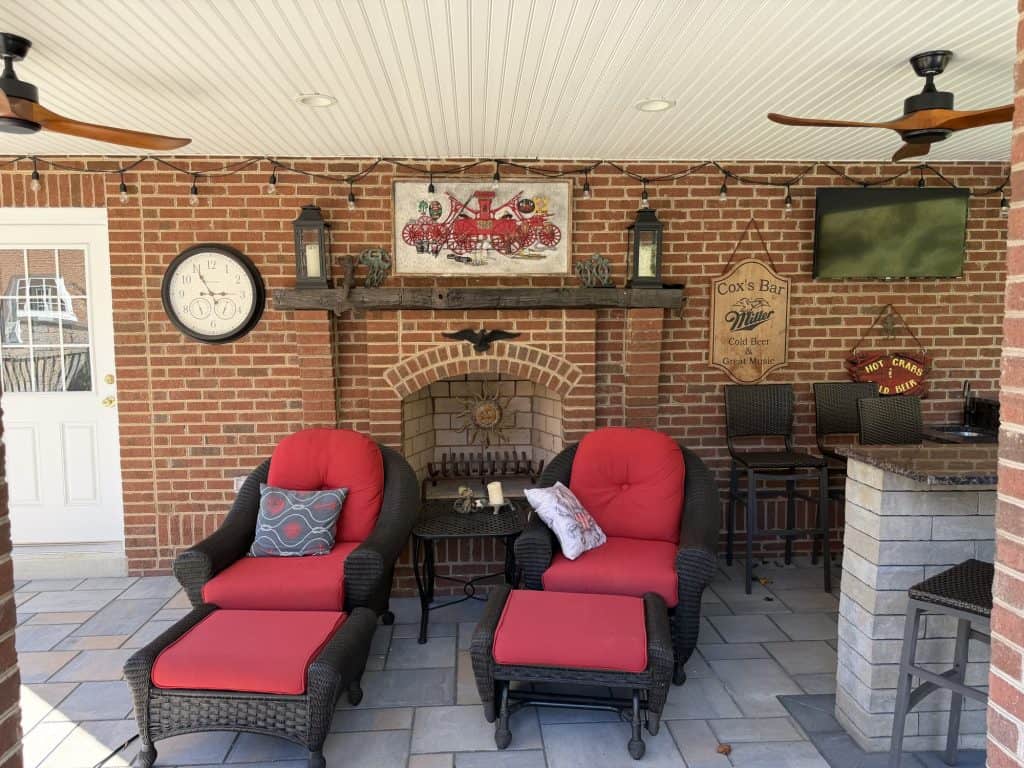 Outdoor living space featuring two red cushioned wicker chairs with matching ottomans, brick fireplace, and decorative wall accents for relaxing outdoor gatherings.