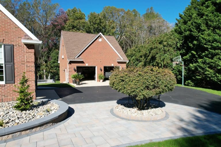 Spacious backyard featuring a basketball court, landscaped garden, and a brick garage with a gable roof under a clear blue sky.